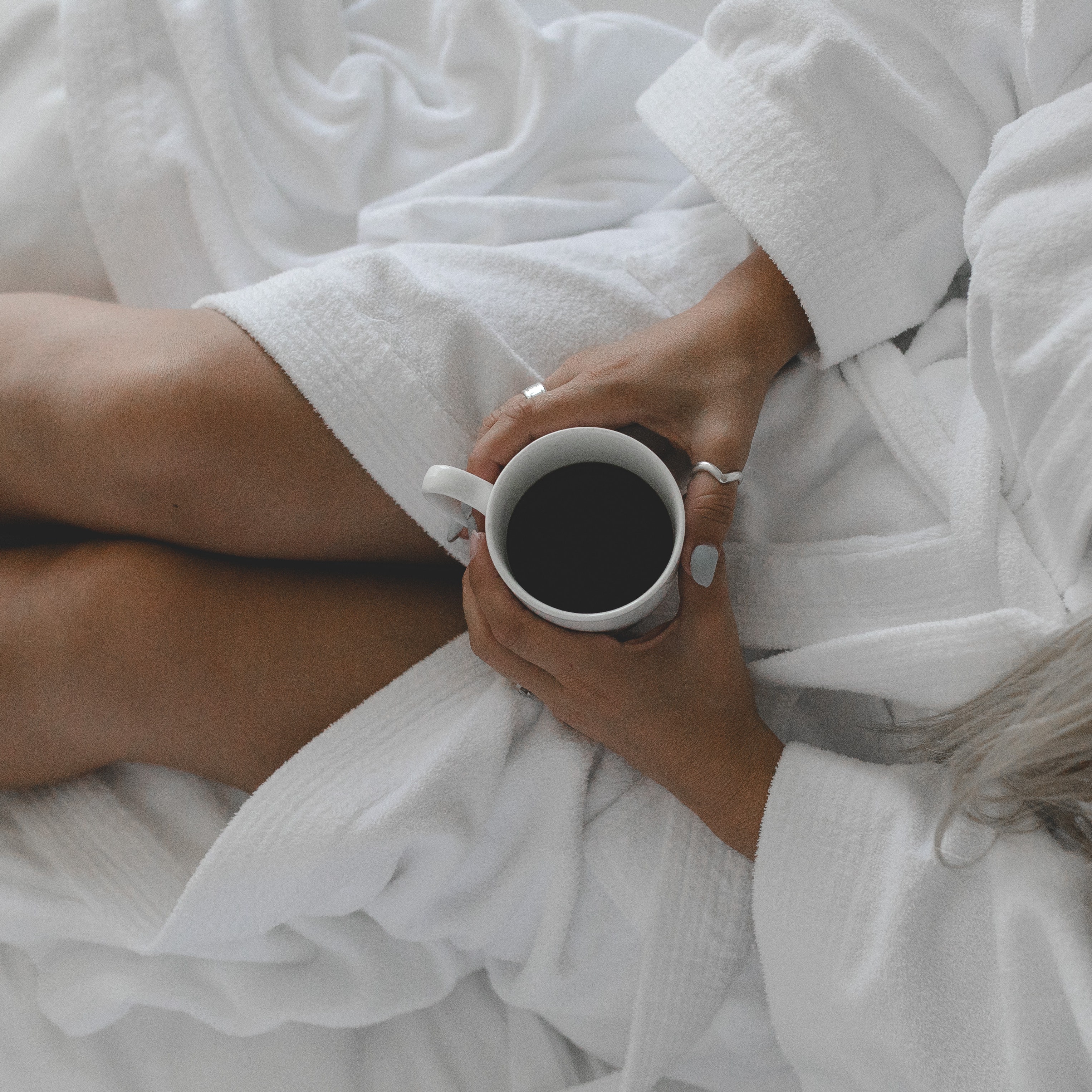 A young tanned woman sitting on a white bed in a white robe holding a coffee cup shot from above.